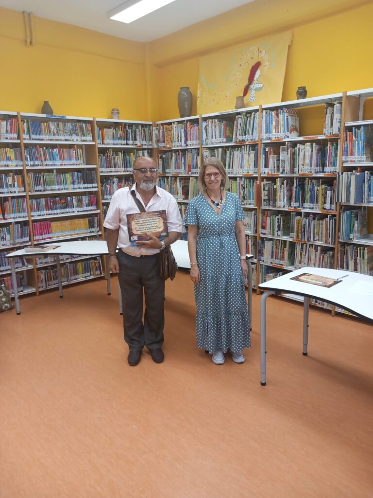 Hombre posa en pie con una mujer y el diploma de ganadoras. Tras ellos, estanterían llenos de libros de la biblioteca.